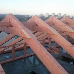 Konstrukcja drewniana dachu z widocznym kominem w tle, w budowie, z perspektywy od dołu