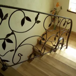 Kuta, metalowa poręcz schodowa z motywem liści, widok z góry na drewniane schody. W tle kremowa ściana i okno. Delikatne światło dzienne.