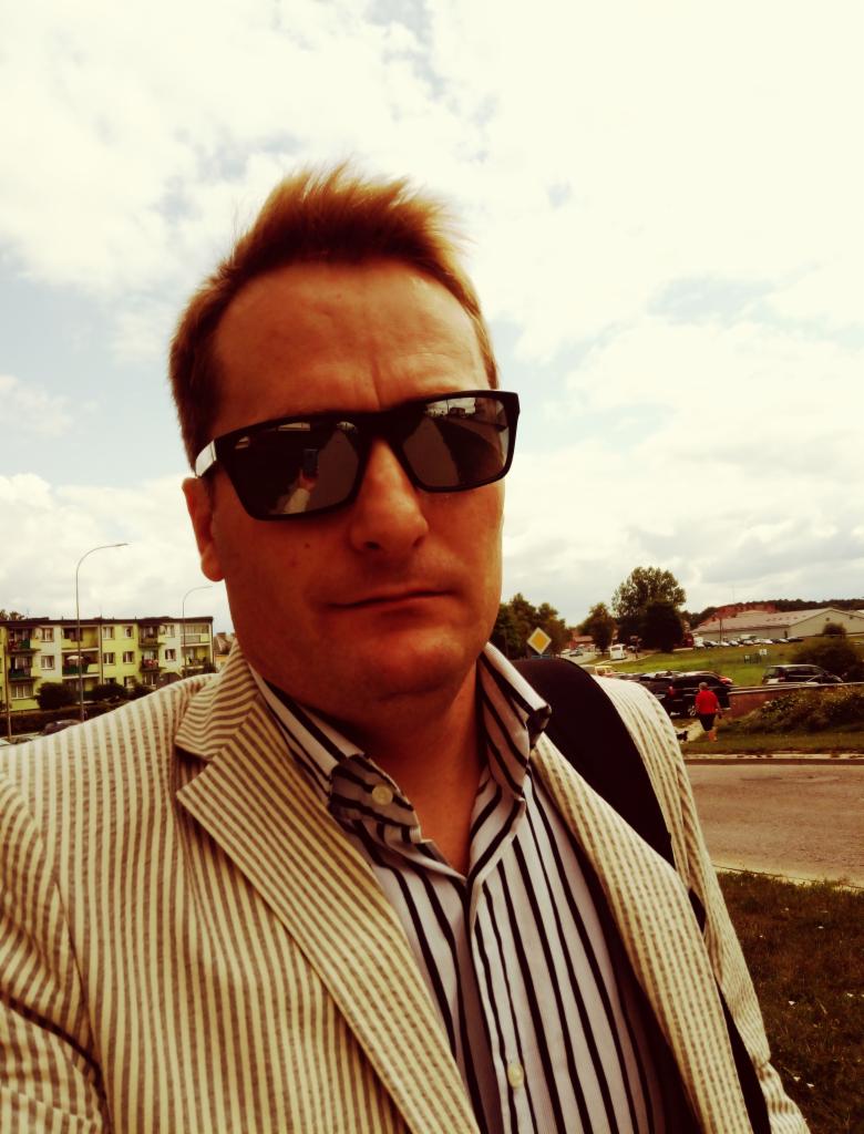 Mężczyzna w okularach przeciwsłonecznych i marynarce w paski, stojący na zewnątrz z budynkami w tle, sugerujący profesjonalny wizerunek.