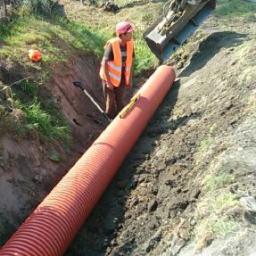REMODOM SPÓŁKA Z OGRANICZONĄ ODPOWIEDZIALNOŚCIĄ - Układanie karbowanej rury drenarskiej w wykopie, z pracownikiem w kamizelce i koparką w tle. Poziomica na rurze.