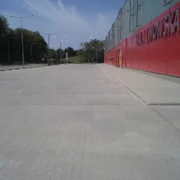 Rozległy parking z kostki brukowej przed budynkiem o czerwonej elewacji z napisem 'HALA LWOWSKA'. Widoczne lampy oświetleniowe i drzewa w tle.