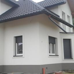 REMODOM SPÓŁKA Z OGRANICZONĄ ODPOWIEDZIALNOŚCIĄ - Dwukondygnacyjny dom z białą elewacją i ciemną dachówką, z widocznymi roletami zewnętrznymi w oknach, z pracownikiem w wejściu, częściowo zakrytym folią ochronną.