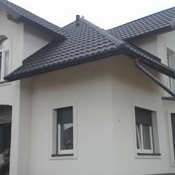 REMODOM SPÓŁKA Z OGRANICZONĄ ODPOWIEDZIALNOŚCIĄ - Nowo wybudowany dom murowany z ciemną dachówką i białymi ścianami, widoczne okna z roletami zewnętrznymi oraz fragment osoby w wejściu.