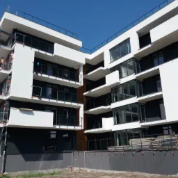 Nowoczesny budynek wielorodzinny w trakcie budowy, widoczne balkony z czarnymi balustradami, białe ściany i czarne elementy elewacji, teren wokół budynku częściowo przygotowany, rury wystające...