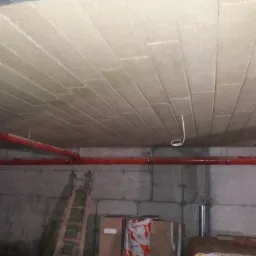 Ocieplenie stropu garażu wełną mineralną w płytach, widoczne rury instalacyjne i drabina oparta o betonową ścianę.