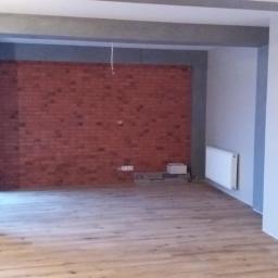 REMODOM SPÓŁKA Z OGRANICZONĄ ODPOWIEDZIALNOŚCIĄ - Surowe wykończenie wnętrza z ceglaną ścianą akcentującą, szarymi elementami architektonicznymi i jasną podłogą z paneli, widoczne niedokończone instalacje elektryczne.