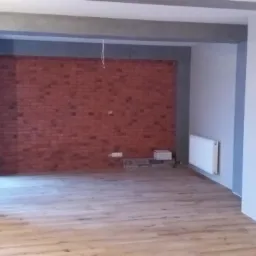 Surowe wykończenie wnętrza z ceglaną ścianą akcentującą, szarymi elementami architektonicznymi i jasną podłogą z paneli, widoczne niedokończone instalacje elektryczne.