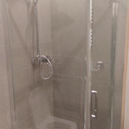 REMODOM SPÓŁKA Z OGRANICZONĄ ODPOWIEDZIALNOŚCIĄ - Szklana kabina prysznicowa z deszczownicą w łazience z szarymi płytkami, widoczny odpływ liniowy w podłodze i toaleta w tle.