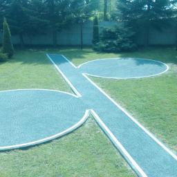 REMODOM SPÓŁKA Z OGRANICZONĄ ODPOWIEDZIALNOŚCIĄ - Oryginalny, dekoracyjny chodnik z kostki brukowej w kształcie dwóch kół połączonych prostą ścieżką, z białym obramowaniem, na tle zielonego trawnika, z tujami i betonowym ogrodzeniem w tle.