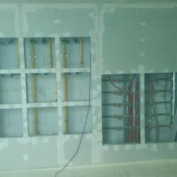 REMODOM SPÓŁKA Z OGRANICZONĄ ODPOWIEDZIALNOŚCIĄ - Ściana z płyt kartonowo-gipsowych w trakcie montażu, widoczne otwory z instalacją elektryczną (żółte rury) i grzewczą (czerwone rury), szpachlowane łączenia płyt.