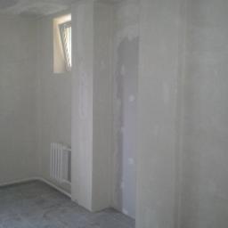 REMODOM SPÓŁKA Z OGRANICZONĄ ODPOWIEDZIALNOŚCIĄ - Wnętrze w trakcie wykończenia: surowe ściany z płyt kartonowo-gipsowych, grzejnik pod oknem, szara podłoga z płytek. Widoczne nierówności i niedoskonałości.