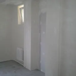 Wnętrze w trakcie wykończenia: surowe ściany z płyt kartonowo-gipsowych, grzejnik pod oknem, szara podłoga z płytek. Widoczne nierówności i niedoskonałości.