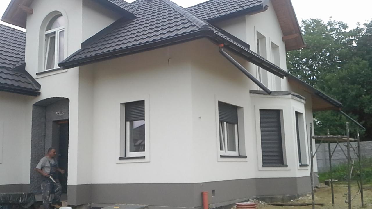 Dwukondygnacyjny dom z białą elewacją i ciemną dachówką, z widocznymi roletami zewnętrznymi w oknach, z pracownikiem w wejściu, częściowo zakrytym folią ochronną.
