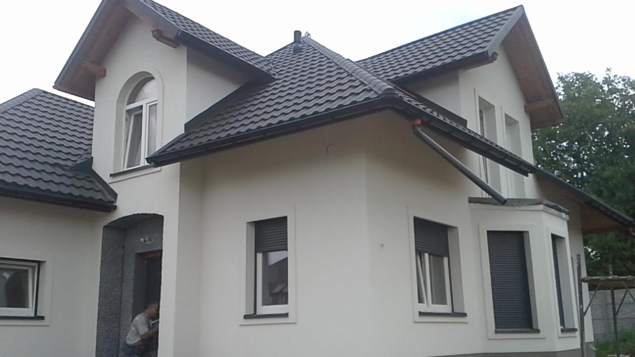 Nowo wybudowany dom murowany z ciemną dachówką i białymi ścianami, widoczne okna z roletami zewnętrznymi oraz fragment osoby w wejściu.