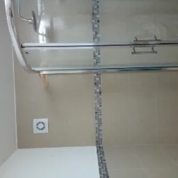 Beżowa łazienka z kabiną prysznicową z przesuwnymi drzwiami, wykończona mozaiką w odcieniach szarości i białym wentylatorem na ścianie.