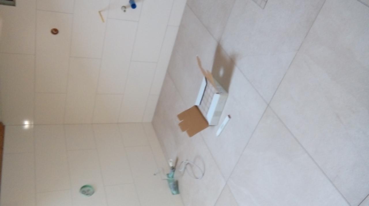 Płytki podłogowe i ścienne w trakcie układania, widoczna otwarta paczka z płytkami, narzędzia glazurnicze i fragment instalacji hydraulicznej.