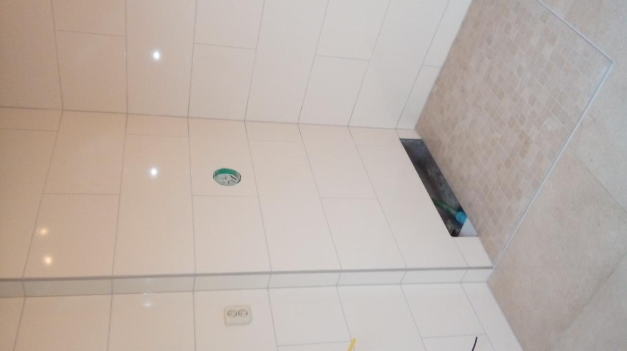 Jasne, prostokątne płytki ścienne w łazience, z widocznym gniazdkiem elektrycznym i fragmentem podłogi wykończonej mozaiką. Nierówności w podłodze wskazują na trwające prace.