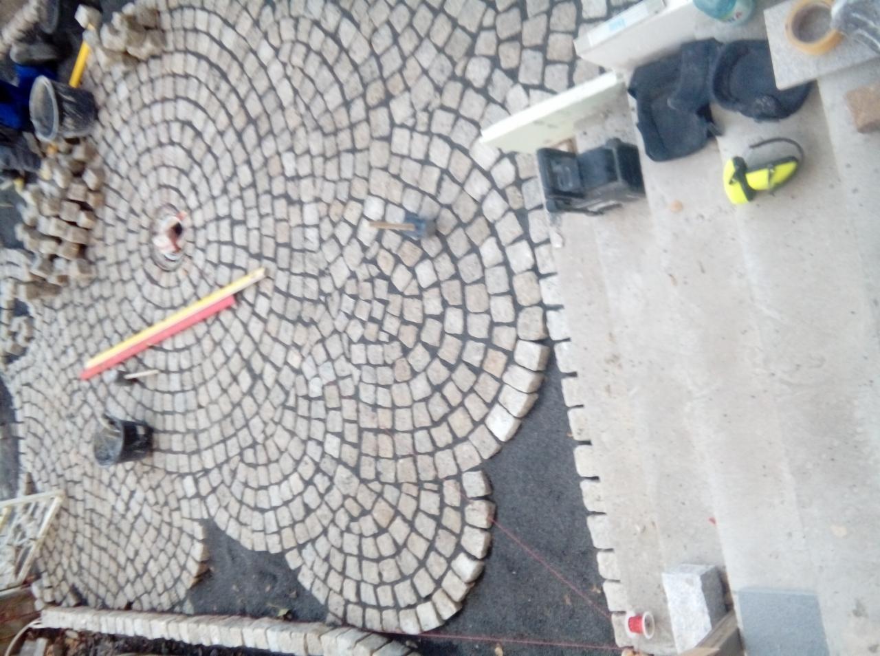 Układanie kostki brukowej w koliste wzory na podwórku, widok z góry, widoczne narzędzia i materiały budowlane, częściowo ułożona nawierzchnia.