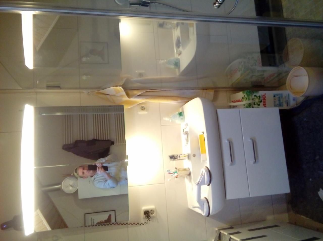 Biała szafka pod umywalką z umywalką i baterią, widoczne akcesoria łazienkowe i odbicie osoby robiącej zdjęcie w lustrze z oświetleniem LED w łazience z białymi płytkami.