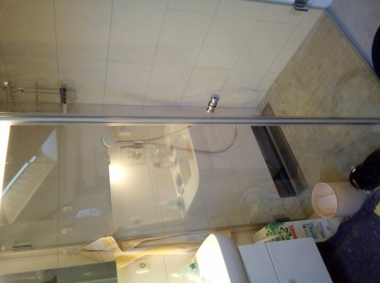 Szklana kabina prysznicowa z widoczną armaturą, białą umywalką i fragmentem płytek podłogowych w łazience, z widocznymi kosmetykami i drobnymi przedmiotami na półce.