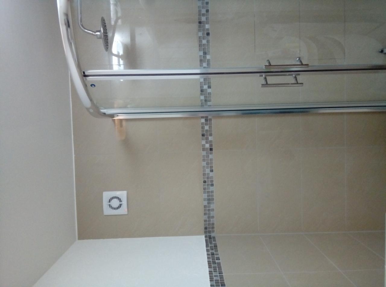 Beżowa łazienka z kabiną prysznicową z przesuwnymi drzwiami, wykończona mozaiką w odcieniach szarości i białym wentylatorem na ścianie.