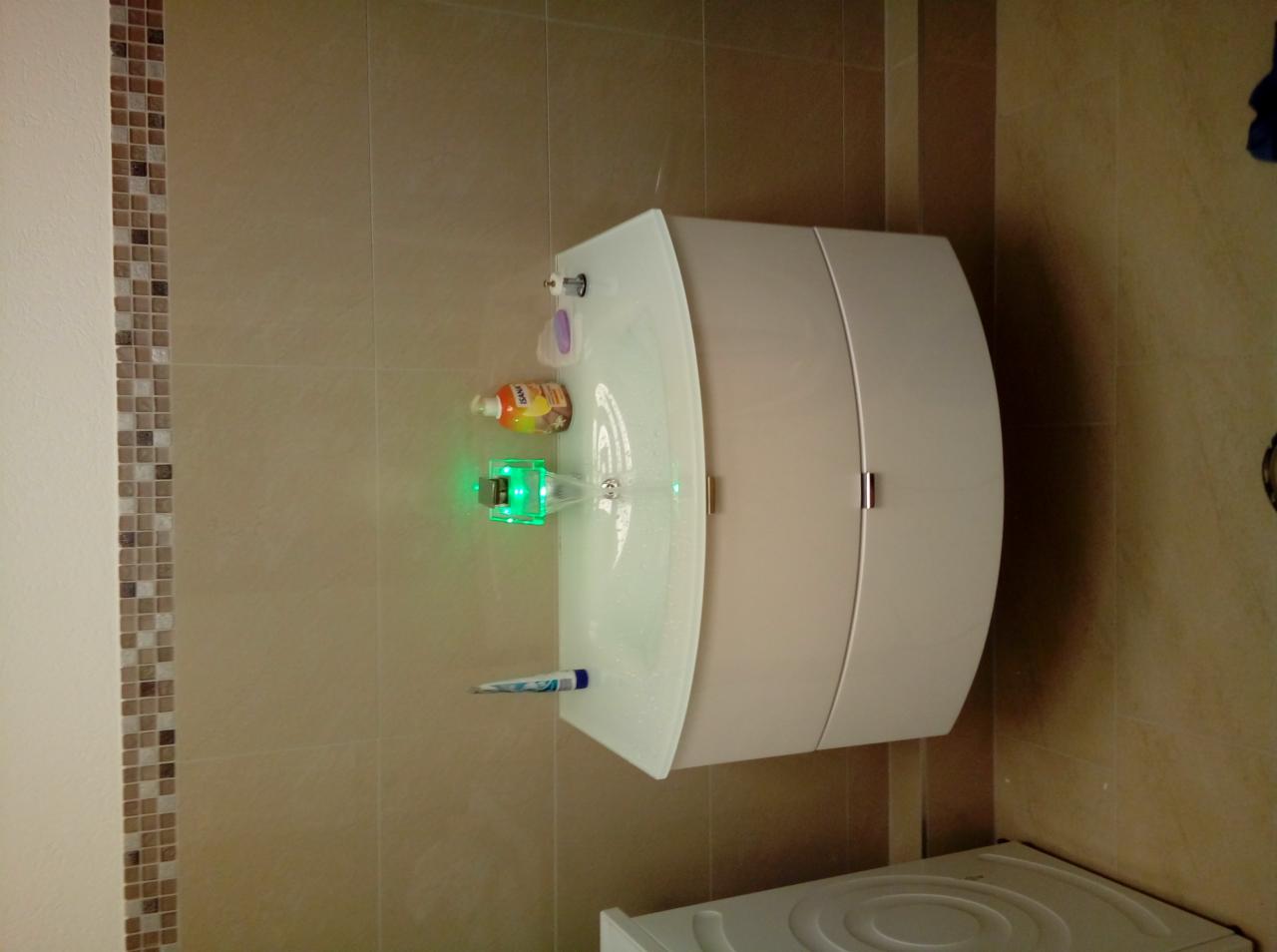 Nowoczesna, podwieszana umywalka z szafką w łazience z beżowymi płytkami, wyposażona w baterię z podświetleniem LED, mydło w płynie i pastę do zębów.