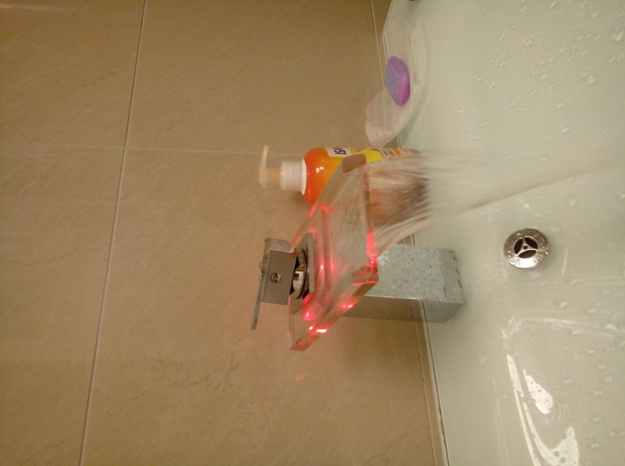 Woda płynąca z nowoczesnej baterii umywalkowej z podświetleniem LED, obok butelka mydła w płynie i mydelniczka z fioletowym mydłem.