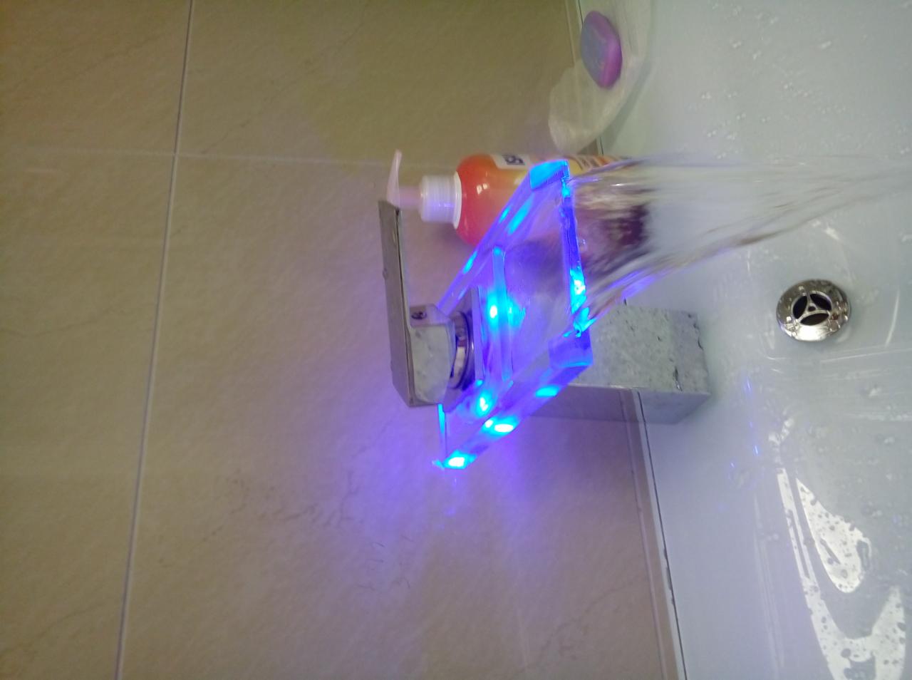 Bateria umywalkowa z podświetleniem LED, z której płynie woda. Obok stoi butelka z mydłem w płynie. Widoczne beżowe płytki ścienne.