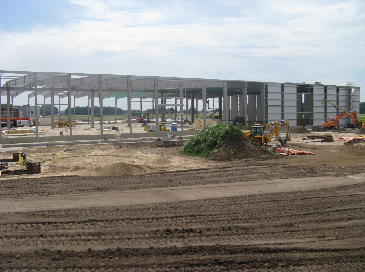 Budowa hali stalowej w trakcie montażu konstrukcji, widoczne maszyny budowlane i teren przygotowany pod dalsze prace, w tle zieleń drzew.