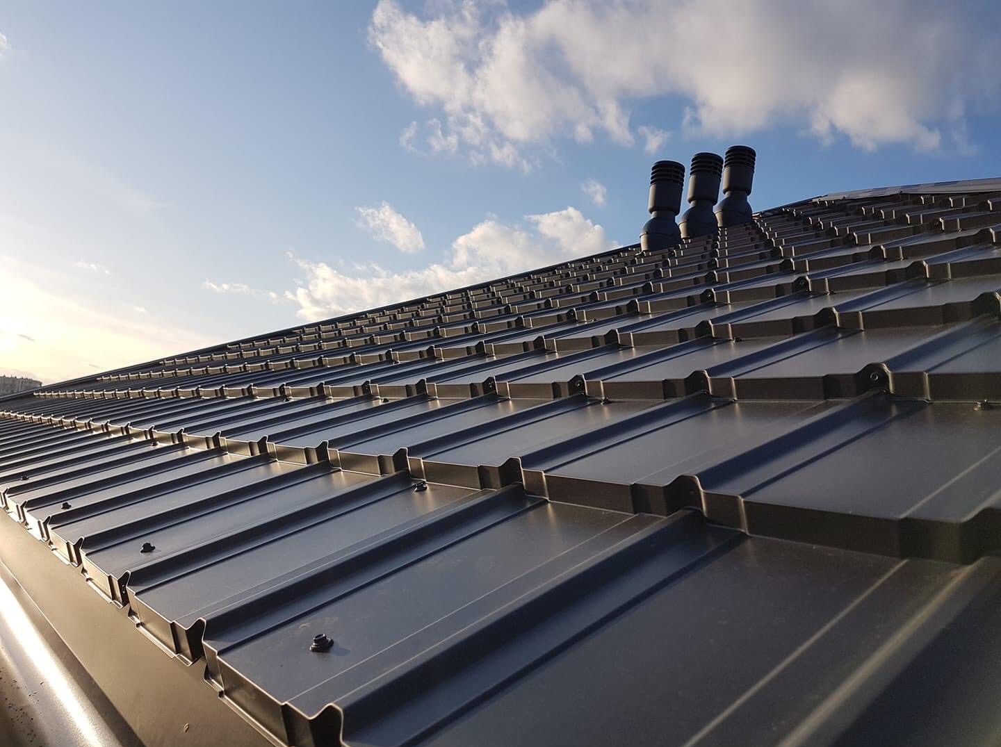 Nowoczesny, grafitowy dach z blachy trapezowej z trzema kominkami wentylacyjnymi na tle błękitnego nieba z chmurami.