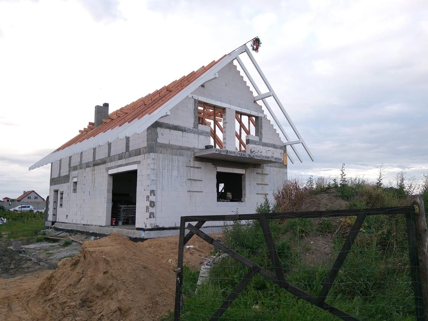 Budowa domu jednorodzinnego w stanie surowym otwartym, widoczna konstrukcja dachu z krokwiami i łatami, ściany z białego bloczka, w tle zachmurzone niebo.