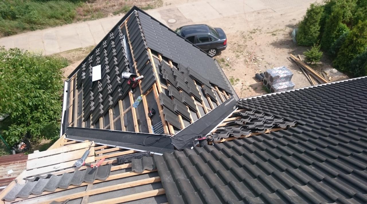 Montaż czarnej dachówki ceramicznej na drewnianej konstrukcji dachu. Widok z góry, narzędzia pracy, częściowo pokryty dach. W tle drzewa i zaparkowany samochód.