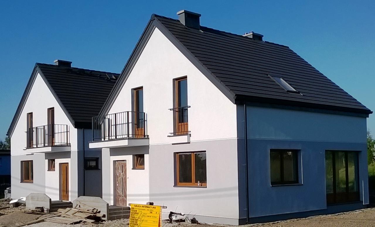 Dwa identyczne, nowoczesne domy jednorodzinne w trakcie budowy, z ciemną dachówką i balkonami z metalową balustradą, na tle błękitnego nieba.