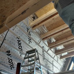 Tomex - Ocieplenie poddasza z folią aluminiową PAROTEC, widoczne drewniane belki i drabina. Prace wykończeniowe na poddaszu, izolacja termiczna w trakcie montażu.