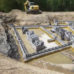 Tomex - Fundamenty domu w trakcie budowy: zalane wodą bloczki betonowe, żółta izolacja TASSU, wykop ziemny i koparka na górze. Widoczny pracownik w szarym ubraniu.