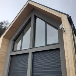 Nowoczesny dom z drewnianą elewacją i dużymi trójkątnymi oknami, zabezpieczonymi szarymi roletami zewnętrznymi.