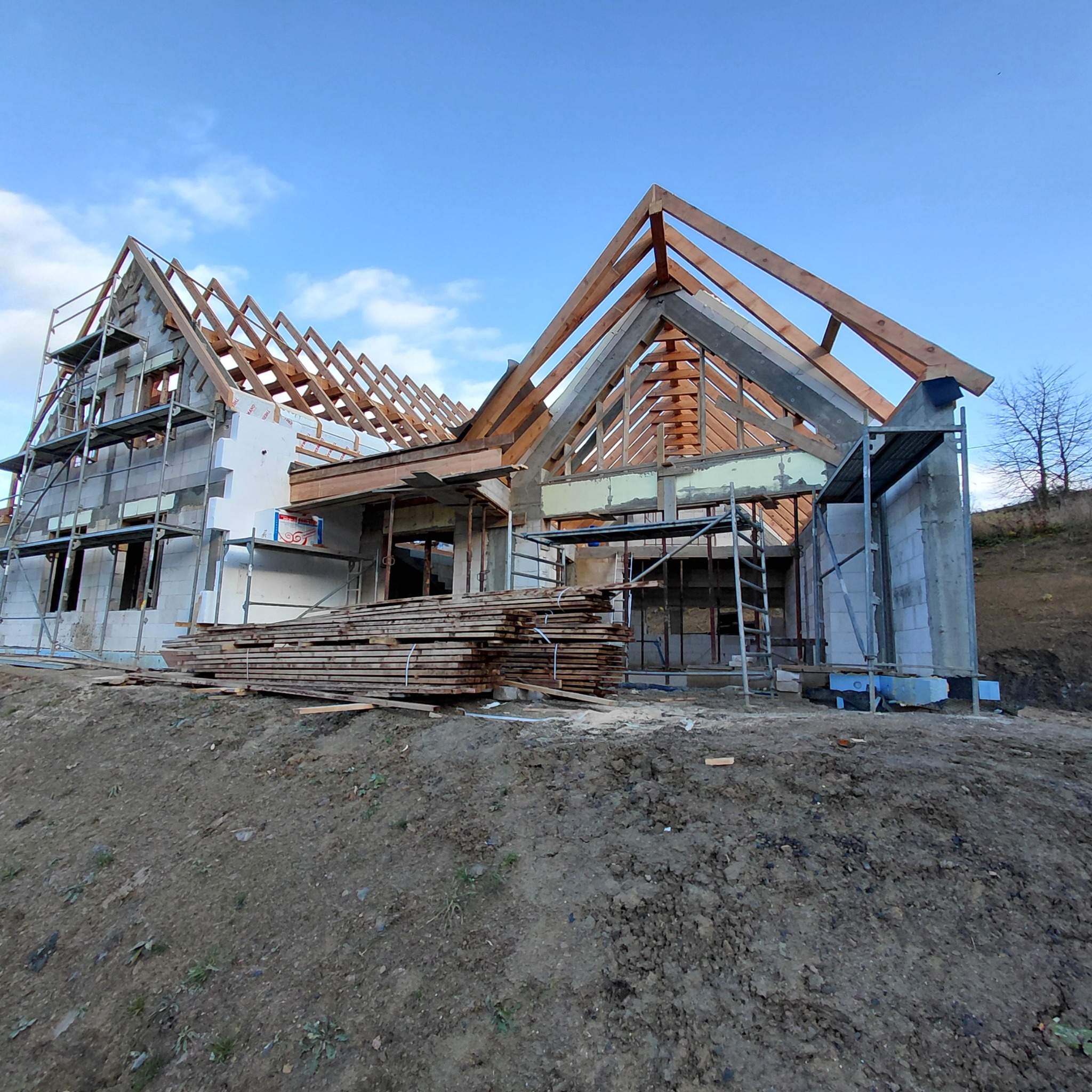 Budowa domu w trakcie realizacji: surowy stan z widocznym drewnianym szkieletem dachu, rusztowaniami i stertą desek na pierwszym planie. Niebo błękitne.