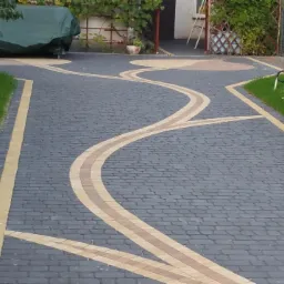 Wzorzysta nawierzchnia z kostki brukowej w odcieniach szarości i beżu, tworząca wijący się wzór na podjeździe, otoczona zielenią trawnika i roślinnością.