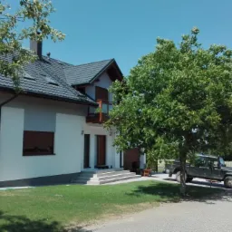 Nowoczesny dom jednorodzinny z szarą dachówką i białymi ścianami, drewnianymi okiennicami i balkonem, z zielonym trawnikiem i podjazdem, na którym stoi terenowy samochód Land Rover Defender.