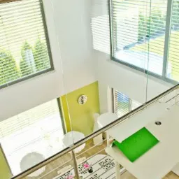 Wnętrze domu z antresolą, widok z góry przez szklaną barierkę. Jasne ściany, duże okna z żaluzjami, białe meble i dywanik z motywem kwiatowym.