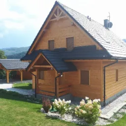 Gotowy, drewniany dom z ciemnym dachem i zadaszeniem na samochód w tle, otoczony zielonym trawnikiem i górskim krajobrazem.