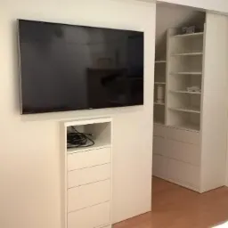 Biała szafa wnękowa z przesuwnymi drzwiami, obok ściana z telewizorem i szafką RTV, minimalistyczny design, widok na okno z żaluzjami.