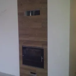 Obudowa kominkowa wykończona płytkami imitującymi drewno, z widocznym wkładem kominkowym i otworami wentylacyjnymi, na tle białej ściany, w trakcie prac wykończeniowych.