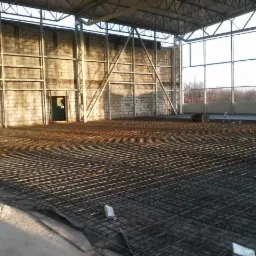 Siatka zbrojeniowa na folii w hali z pustaków, przygotowana pod wylewkę. W tle ściana, okna i elementy konstrukcji dachu. Widoczne narzędzia pomiarowe.