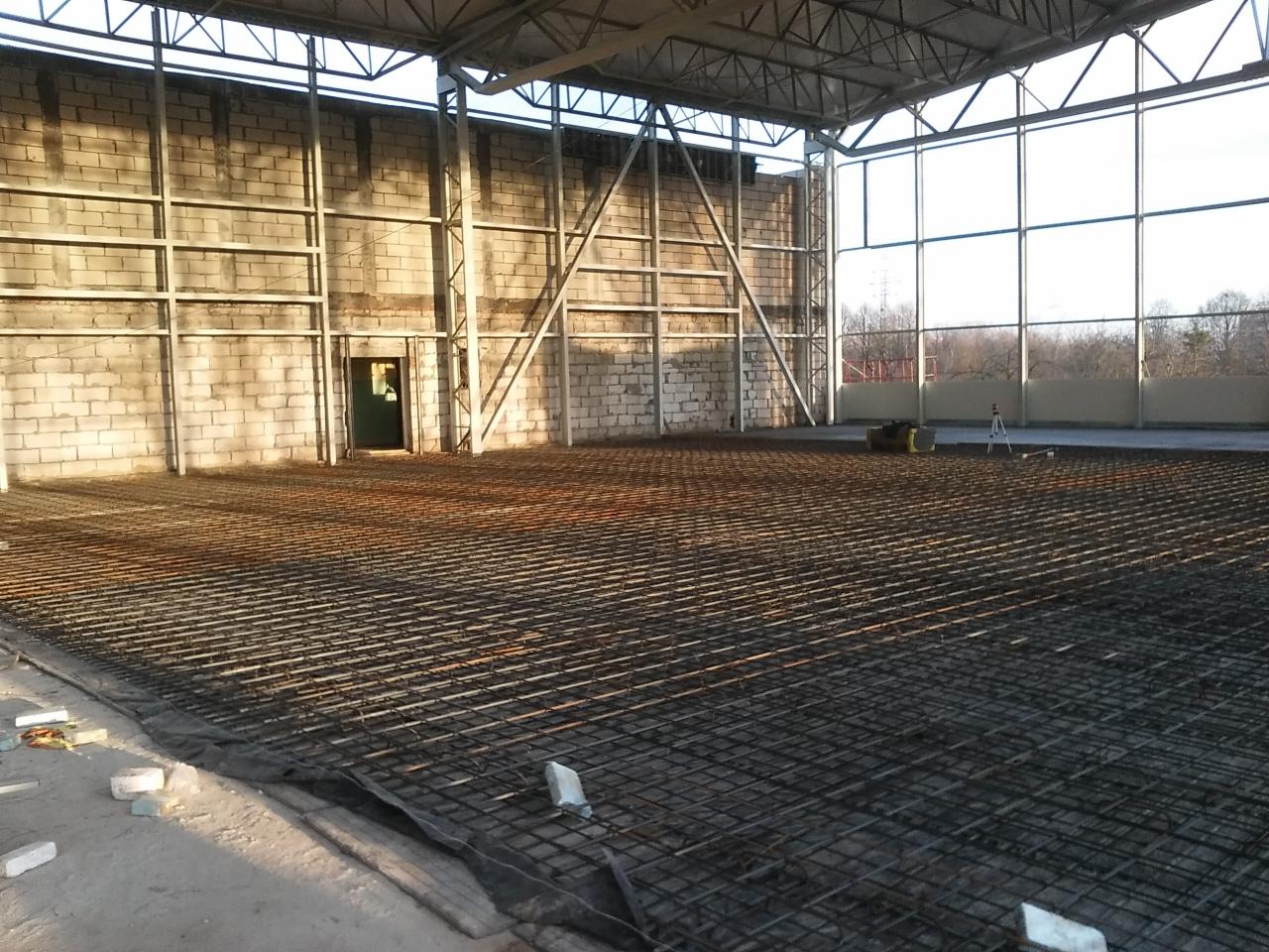 Siatka zbrojeniowa na folii w hali z pustaków, przygotowana pod wylewkę. W tle ściana, okna i elementy konstrukcji dachu. Widoczne narzędzia pomiarowe.