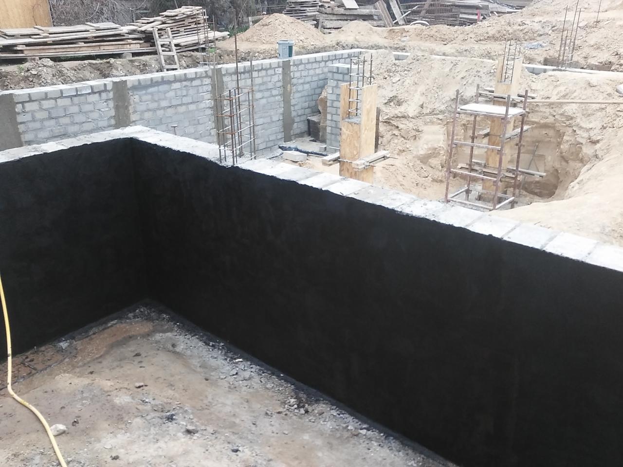 Zabezpieczone fundamenty budynku w trakcie budowy. Widoczne bloczki betonowe, stalowe zbrojenie i hydroizolacja. Na drugim planie wykop i drewniane elementy szalunku.