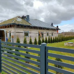 Montaż antracytowej dachówki na domu jednorodzinnym; widoczni dekarze pracujący na dachu, rusztowania, częściowo ułożona dachówka oraz ogrodzenie posesji z tujami.