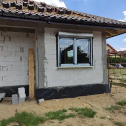 Nowe okno z roletą zewnętrzną zamontowane w surowym budynku z białego bloczka, widoczne elementy konstrukcyjne dachu i rusztowanie w tle.