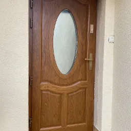 Brązowe drzwi wejściowe z owalnym przeszkleniem i złotą klamką, zamontowane w ościeżnicy z beżowym tynkiem strukturalnym, widoczne zawiasy i próg z płytek.