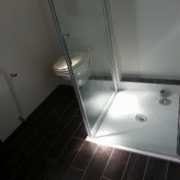 Kabina prysznicowa z brodzikiem, widok z góry, ciemna podłoga imitująca drewno, białe płytki na ścianach, toaleta w tle.